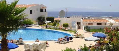 A pool area with lounge chairs and umbrellas, surrounded by greenery and a view of the sea.