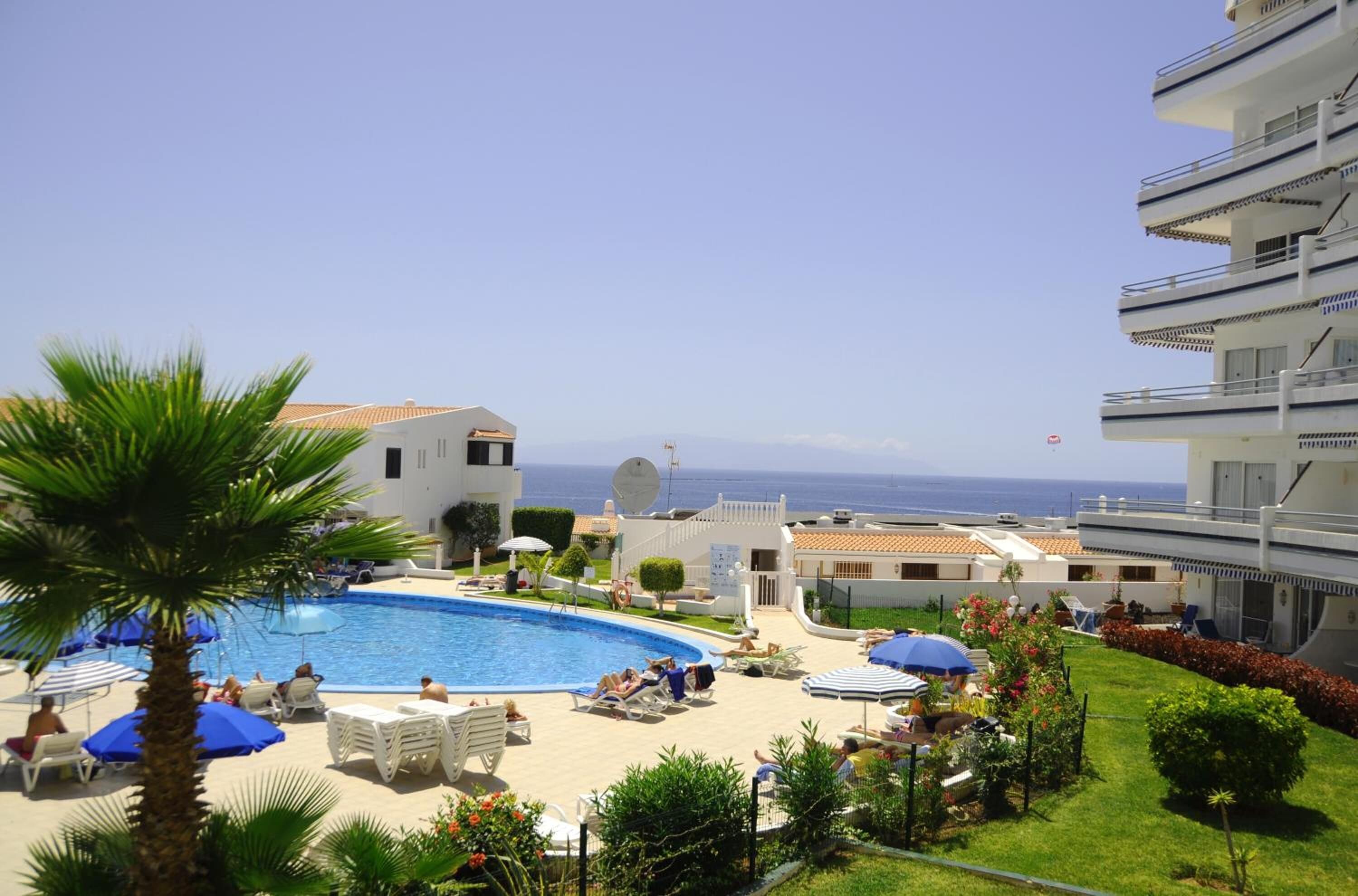 A pool area with lounge chairs and umbrellas, surrounded by greenery and buildings with balconies overlooking the sea.