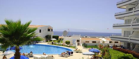 A pool area with lounge chairs and umbrellas, surrounded by greenery and buildings with balconies overlooking the sea.