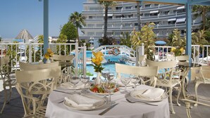 An outdoor dining area with white tables and chairs, a pool in the background, and a multi-story building with balconies. - Hovima Atlantis (Adeje)