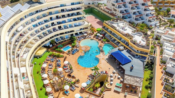 Een hotel met meerdere verdiepingen, een zwembad in het midden, buitenliggende ligbedden en een zonneterras.
