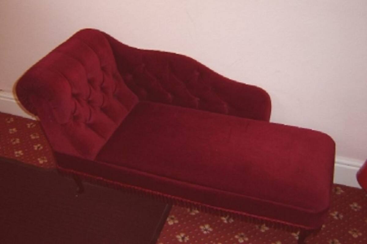 A red velvet lounge chair with a tufted backrest and armrests, positioned against a plain wall with a patterned carpet.
