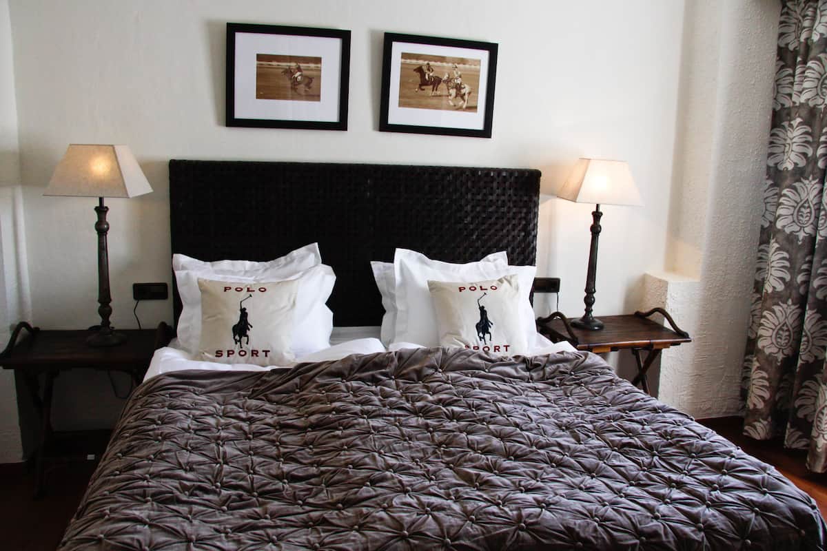 Una cama bien hecha con una colcha oscura y capitonada y dos almohadas con el texto 'POLO SPORT'. Encima de la cama hay dos cuadros enmarcados en la pared, y dos lámparas de noche a cada lado.