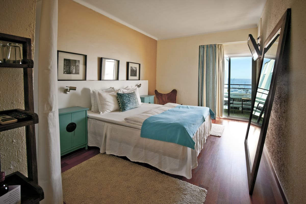 Habitación de hotel con cama, mesita de noche, un armario verde, ventana con cortinas y vista al océano.