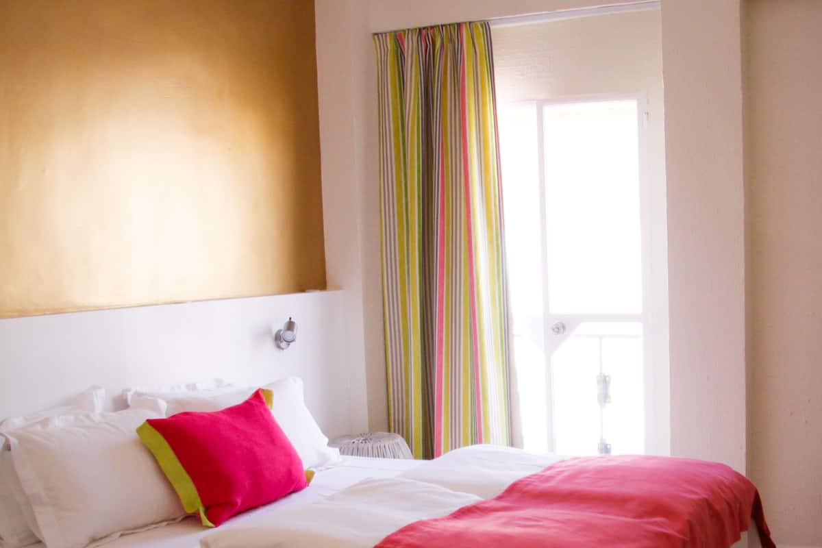 Una habitación de hotel con una cama, ropa de cama blanca, una manta roja y una cortina a rayas coloridas.