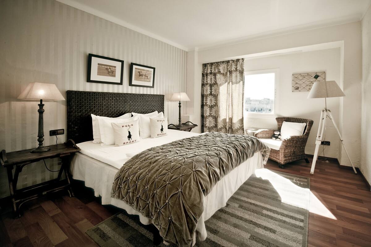 Habitación de hotel con una cama grande, dos mesitas de noche, una silla, una ventana con cortinas y vistas a la ciudad.