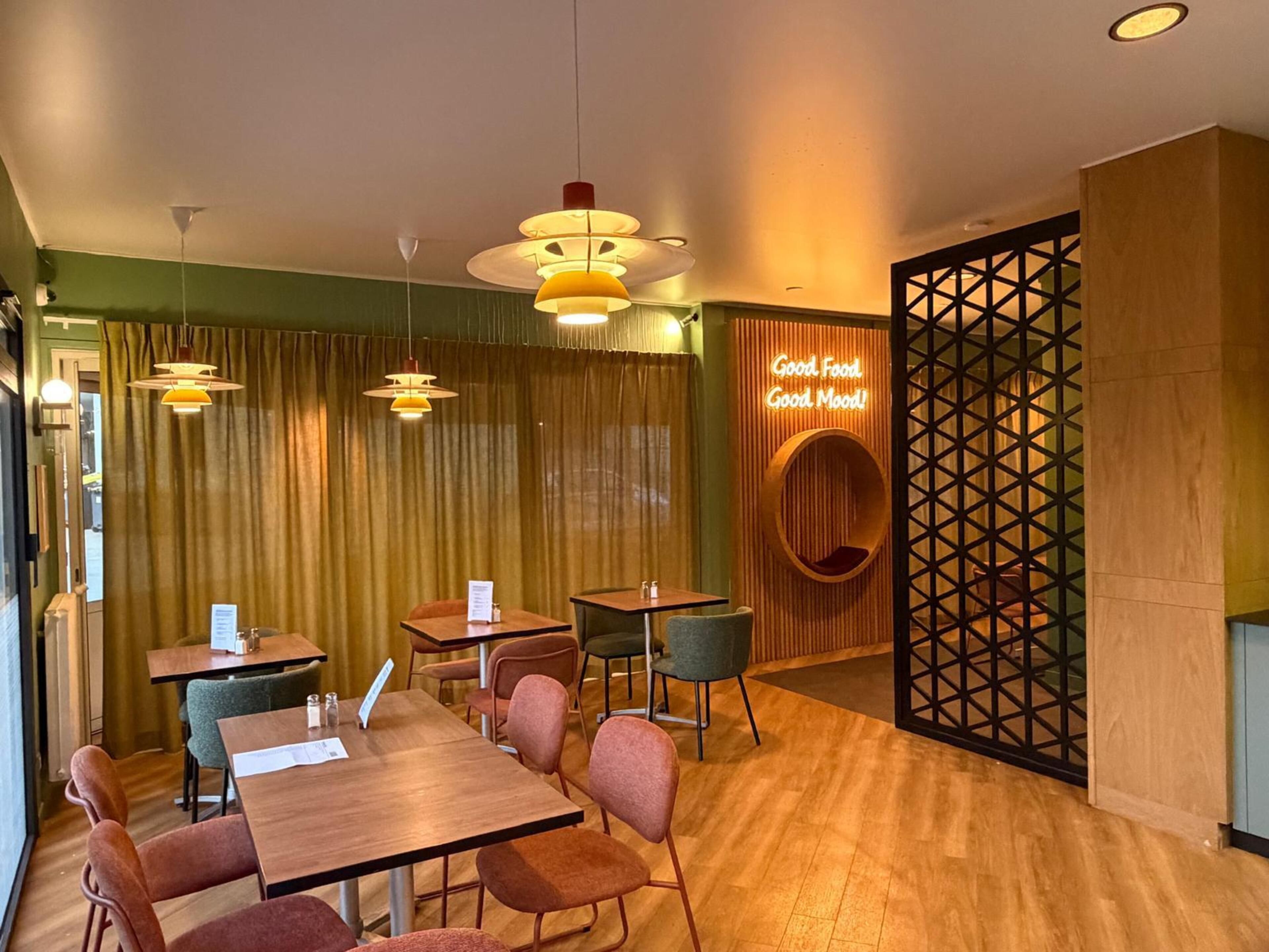 A restaurant interior with wooden tables, cushioned chairs, pendant lighting, and a decorative wall with a circular sign.