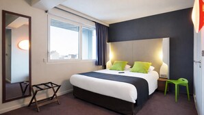 Ein modernes Hotelzimmer mit einem großen Bett, einem Nachttisch, einem kleinen Hocker, einem Spiegel und einem Fenster mit Vorhängen.