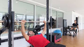 A gym with a man performing a bench press, a woman sitting on a bench, and exercise equipment.