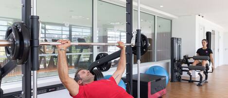 Un gimnasio con un hombre haciendo press de banca, una mujer sentada en un banco y equipo de ejercicio.