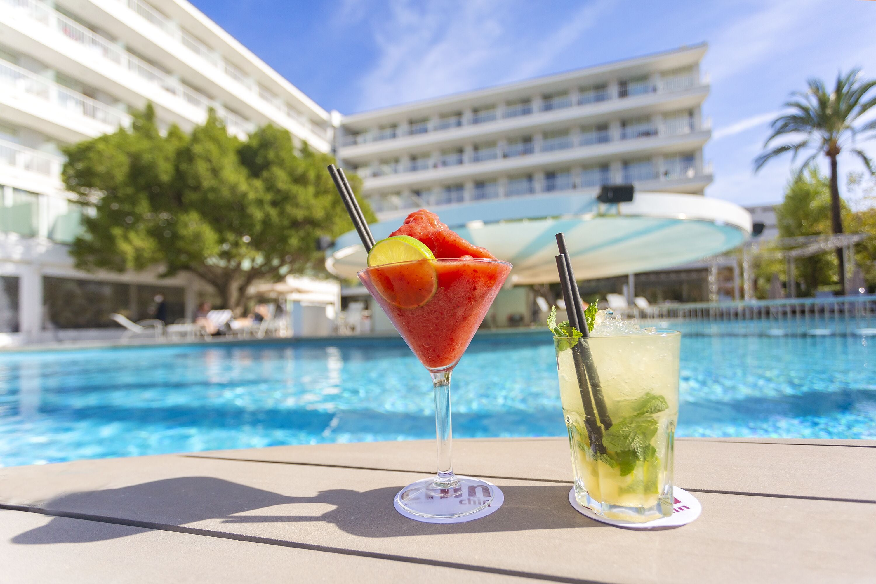 Une scène au bord de la piscine, avec un verre à la main et un cocktail servi dans un autre verre, et un bâtiment d’hôtel en arrière-plan.
