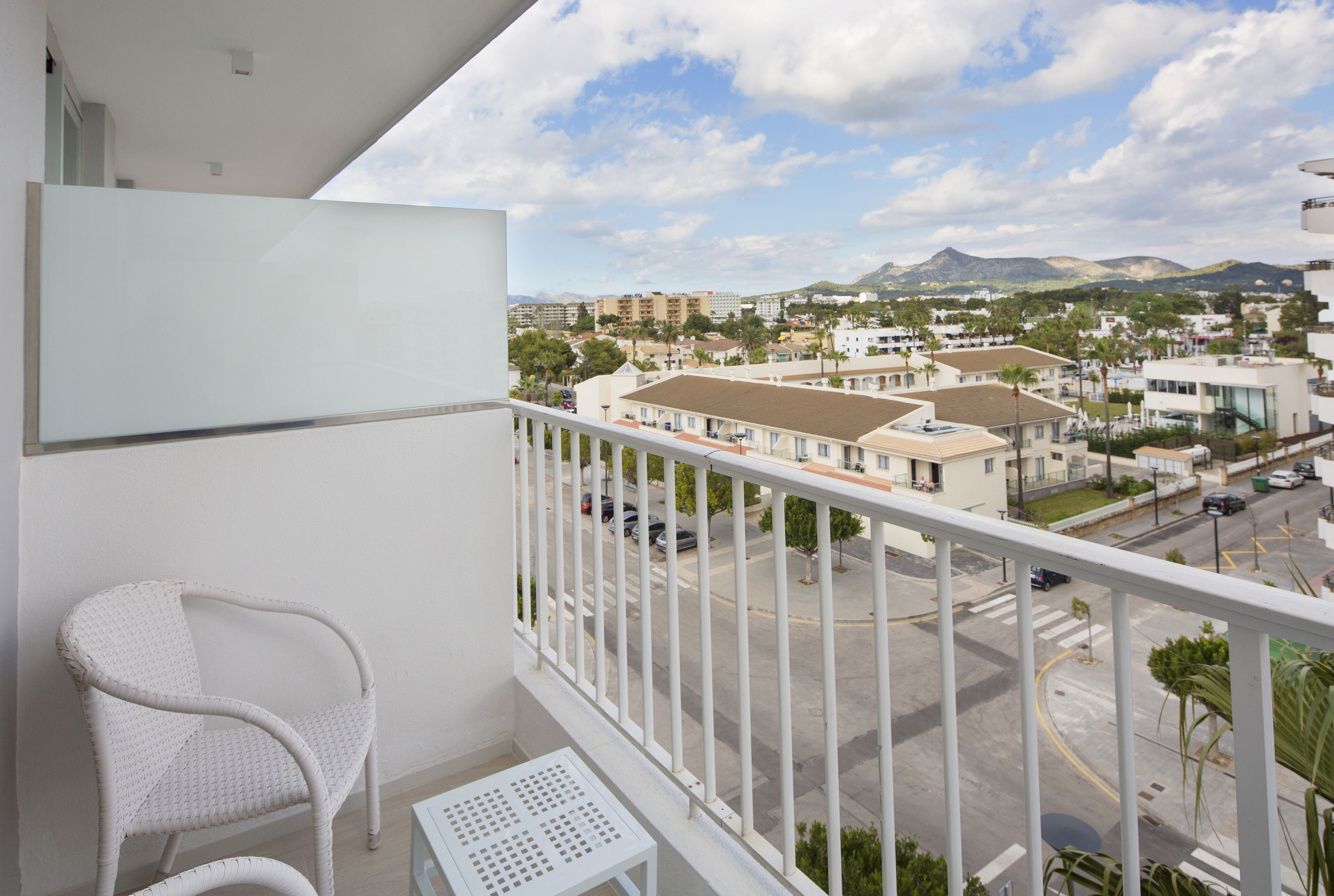A balcony with a white chair and a small table, overlooking a street with buildings and a mountain in the distance.