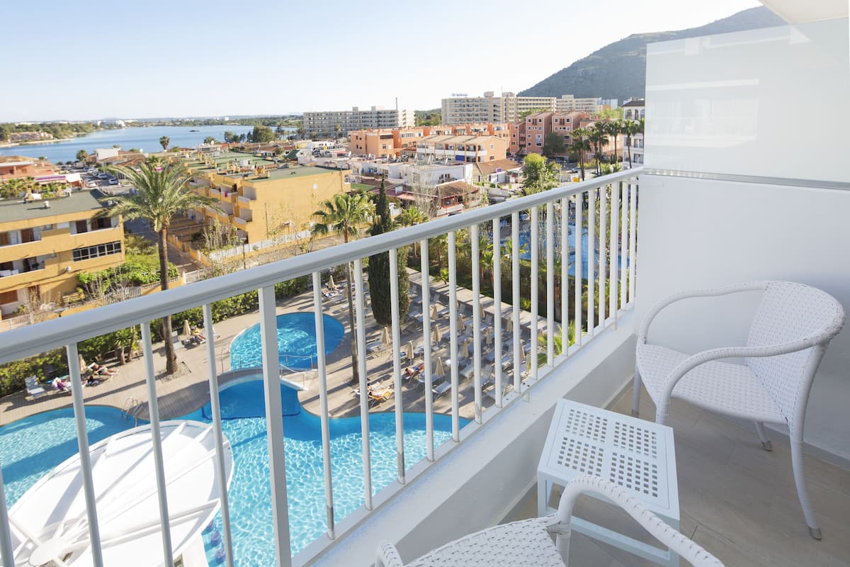 A balcony with white wicker furniture overlooking a swimming pool area with multiple pools and lounge chairs.
