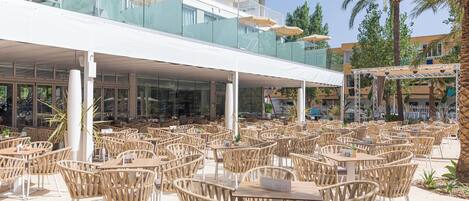Un área de café al aire libre con sillas y mesas de mimbre, una barandilla de vidrio y sombrillas.