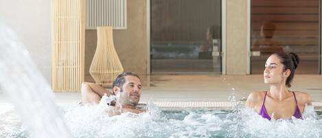 Una pareja se relaja en un jacuzzi con vistas a un edificio moderno y una lámpara de bambú al fondo.