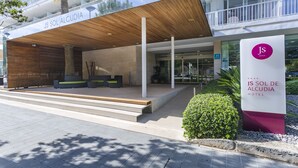 A modern hotel entrance with a covered outdoor seating area, a palm tree, and a sign for JS Sol de Alcudia Hotel. - Hotel JS Sol de Alcudia (Alcúdia)