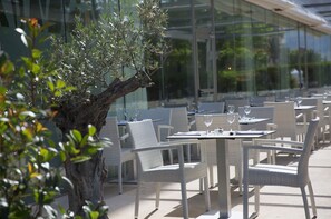 An outdoor dining area with wicker chairs and tables, a large tree, and a glass building in the background. - Hotel JS Sol de Alcudia (Alcúdia)