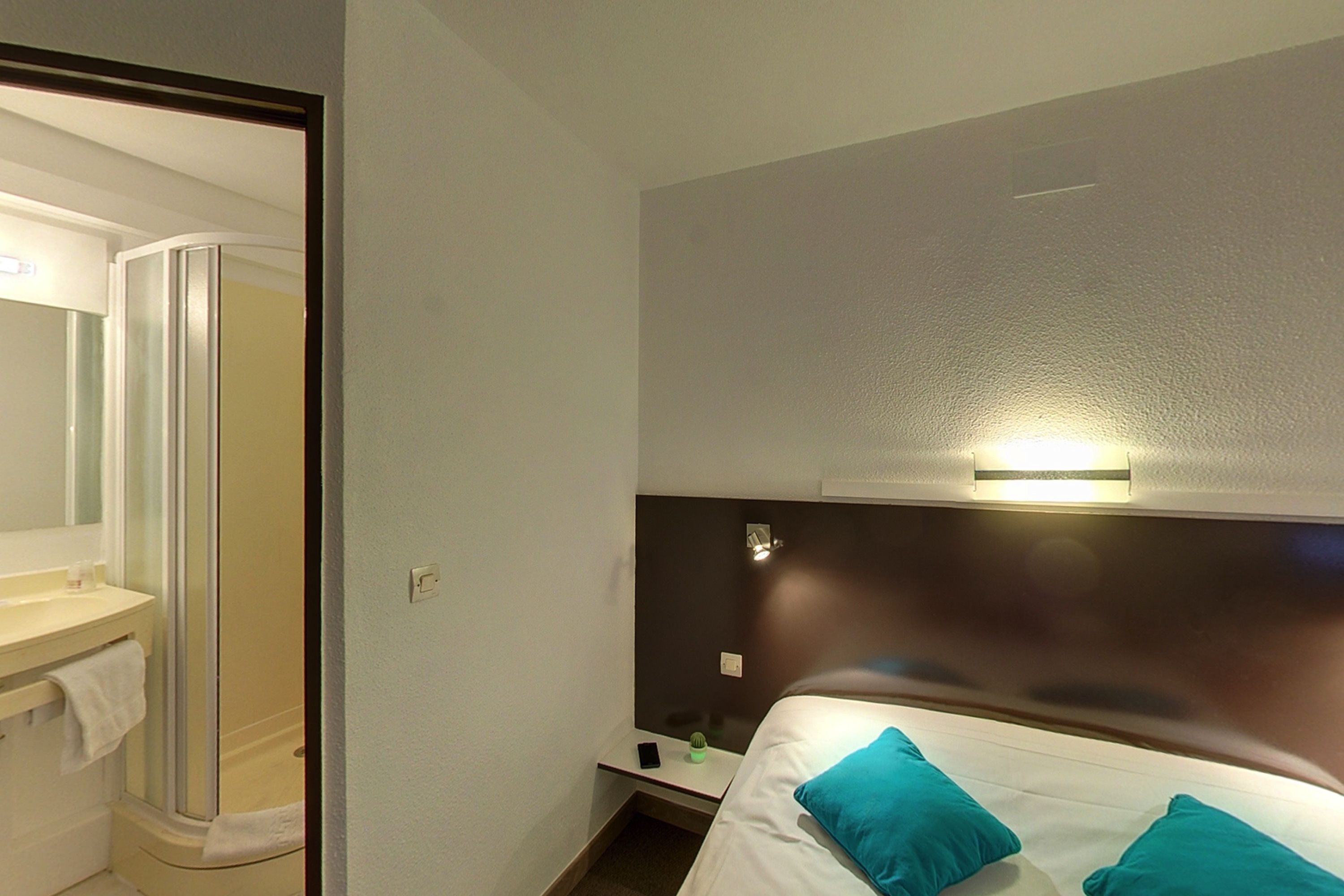 A hotel room with a bed, a nightstand, a small table, a mirror, and a shower area.