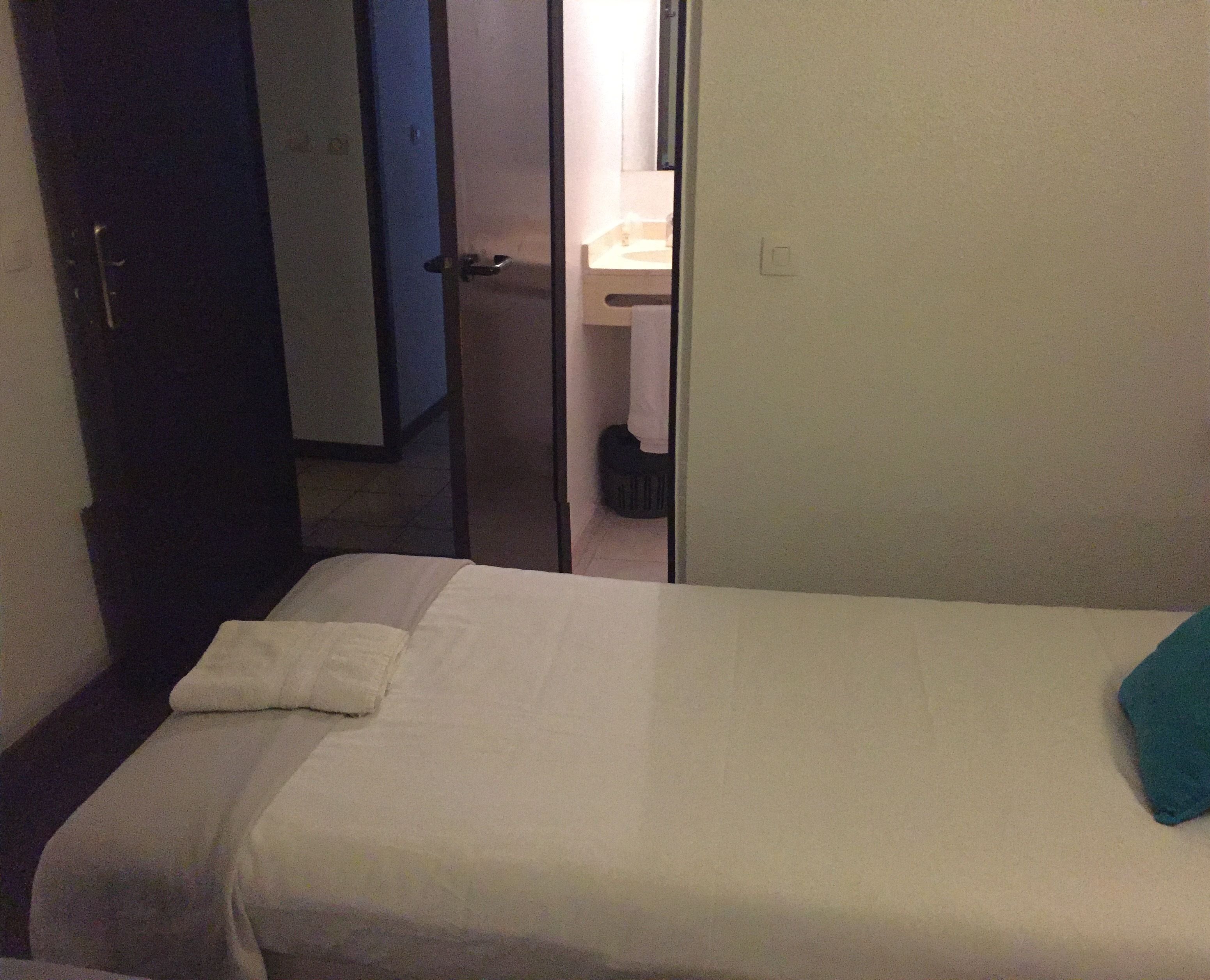 A small, dimly lit room with a single bed, a door leading to a bathroom, and a visible trash bin.