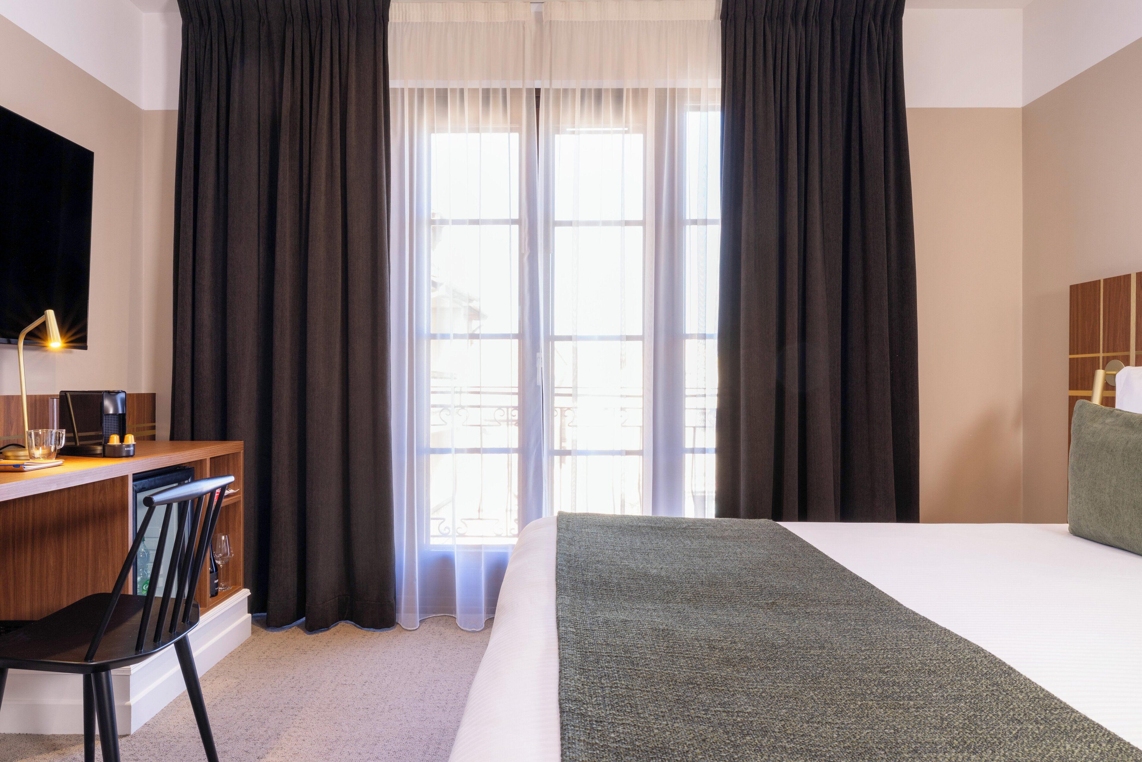 A hotel room with a bed, a desk with a chair, a television mounted on the wall, and a window with curtains.