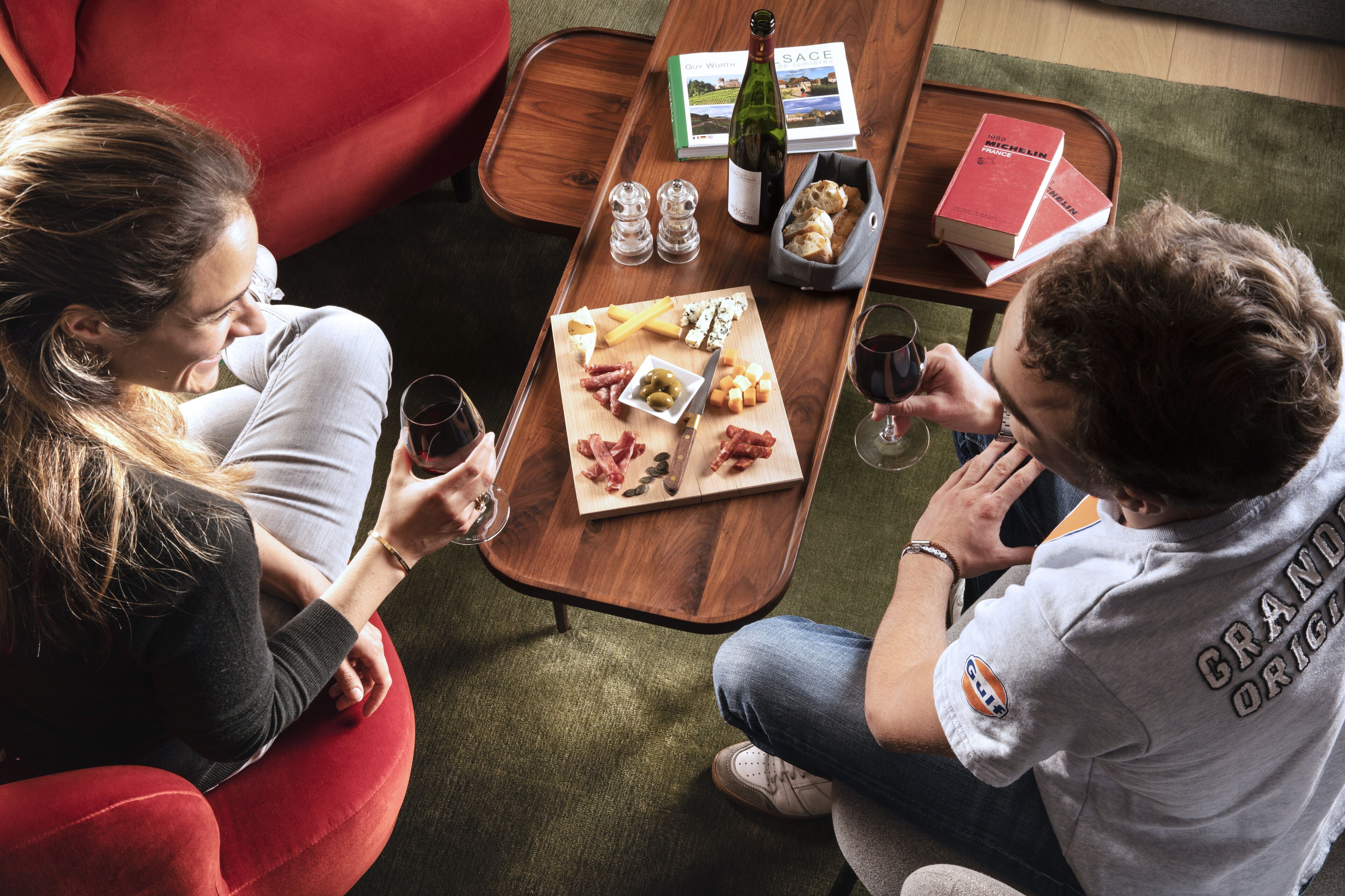 A couple seated at a wooden table with a variety of snacks and drinks, surrounded by books and a bottle of wine.