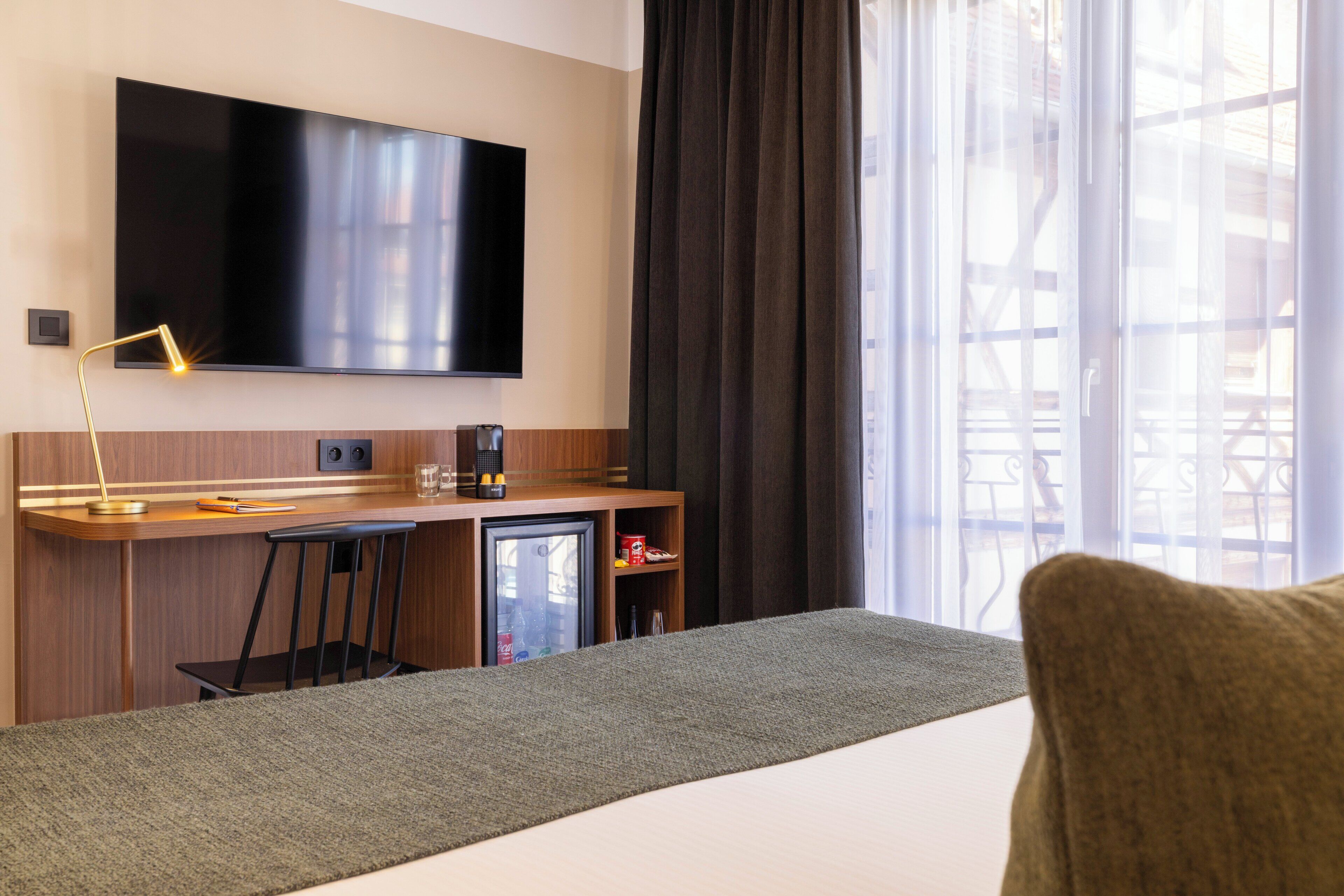 A hotel room with a bed, a desk with a chair, a television, a mini-fridge, and a window with curtains.