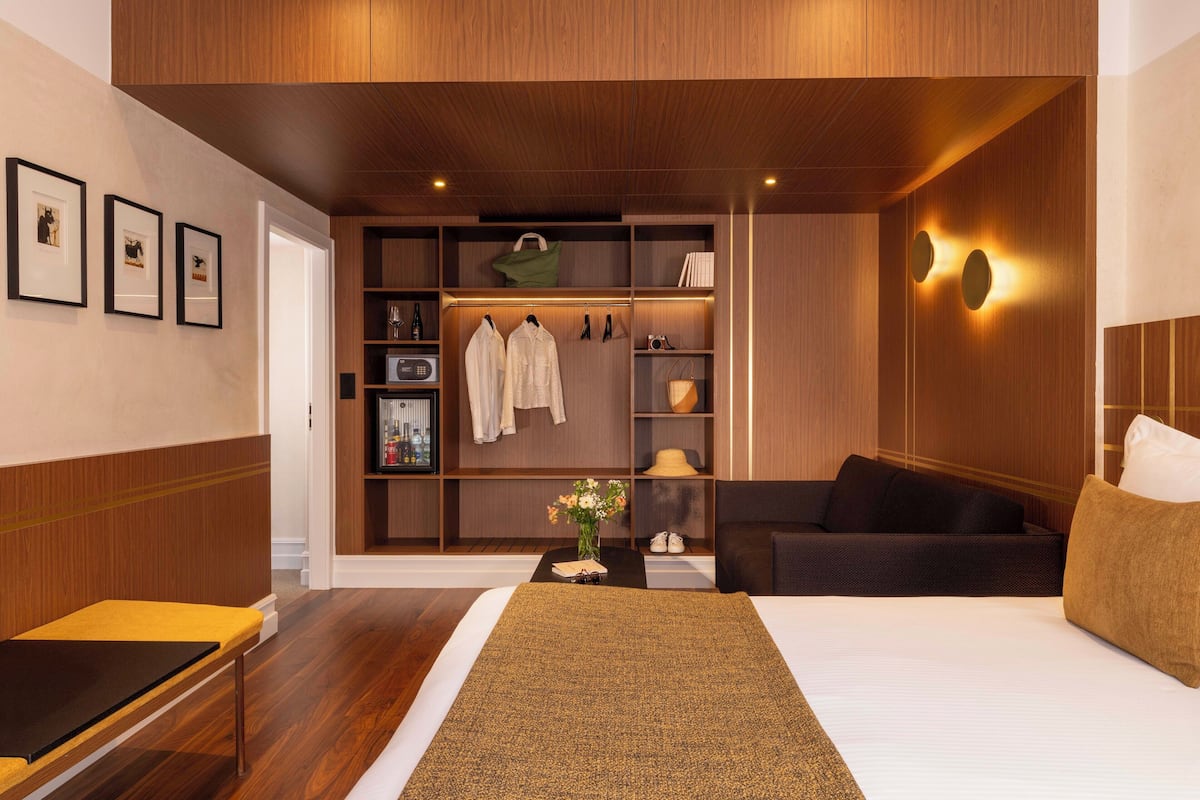 Ein modernes Hotelzimmer mit einem großen Bett, einer Sitzecke mit einer Couch, einem kleinen Tisch mit Blumenvase, einem Kleiderständer mit Kleidung und einem eingebauten Kleiderschrank.