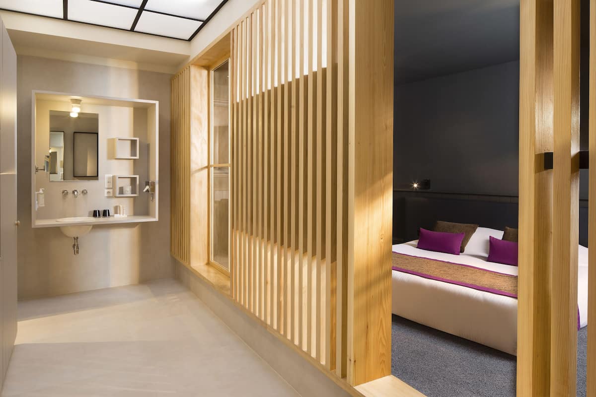 Ein modernes Hotelzimmer mit einer Holzwand, einem großen Bett mit violetten Kissen und einem Badezimmer mit Spiegel und Waschbecken.