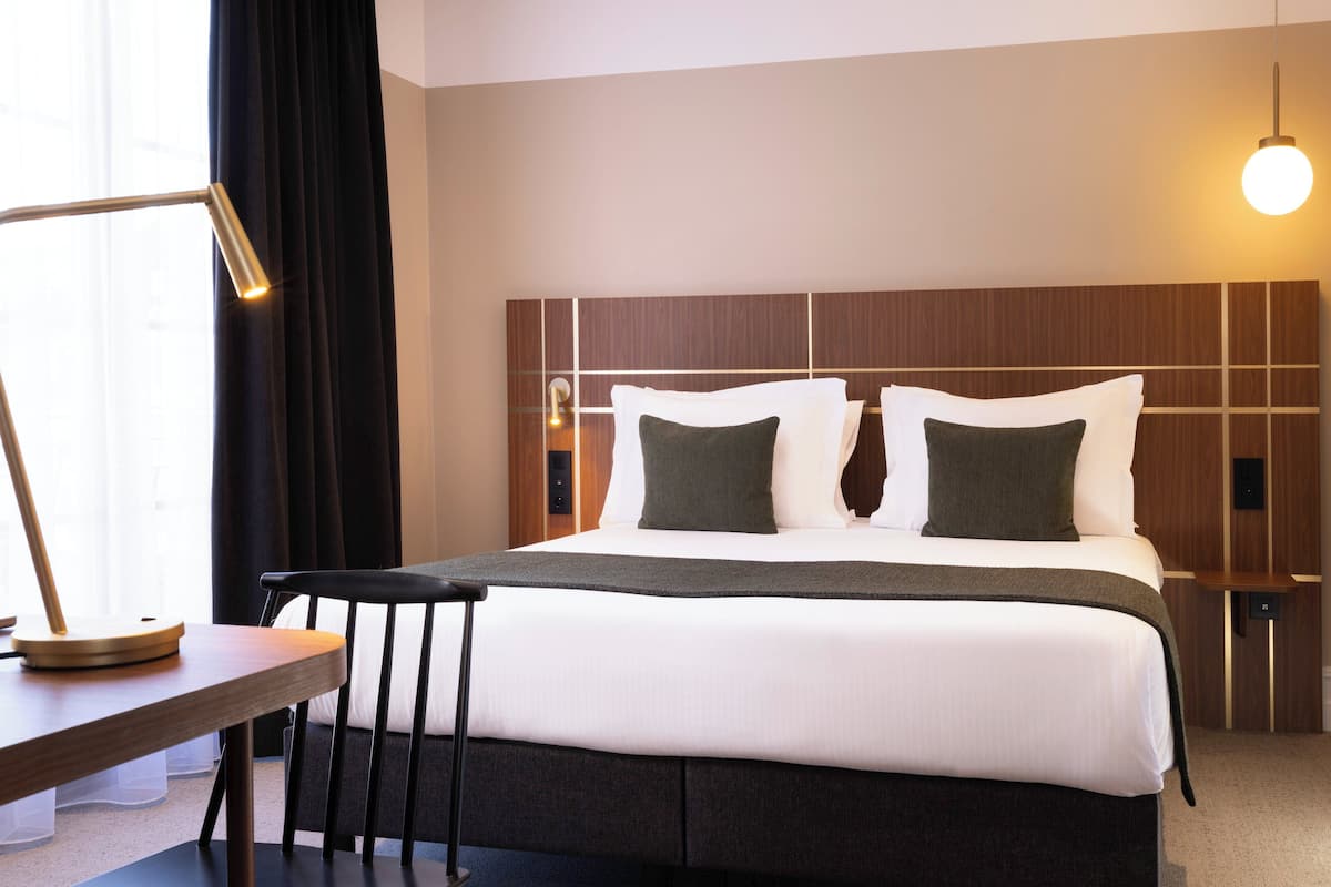 Ein modernes Hotelzimmer mit einem ordentlich bezogenen Bett, einem Schreibtisch mit Lampe und Blick auf die Stadt durch große Fenster.
