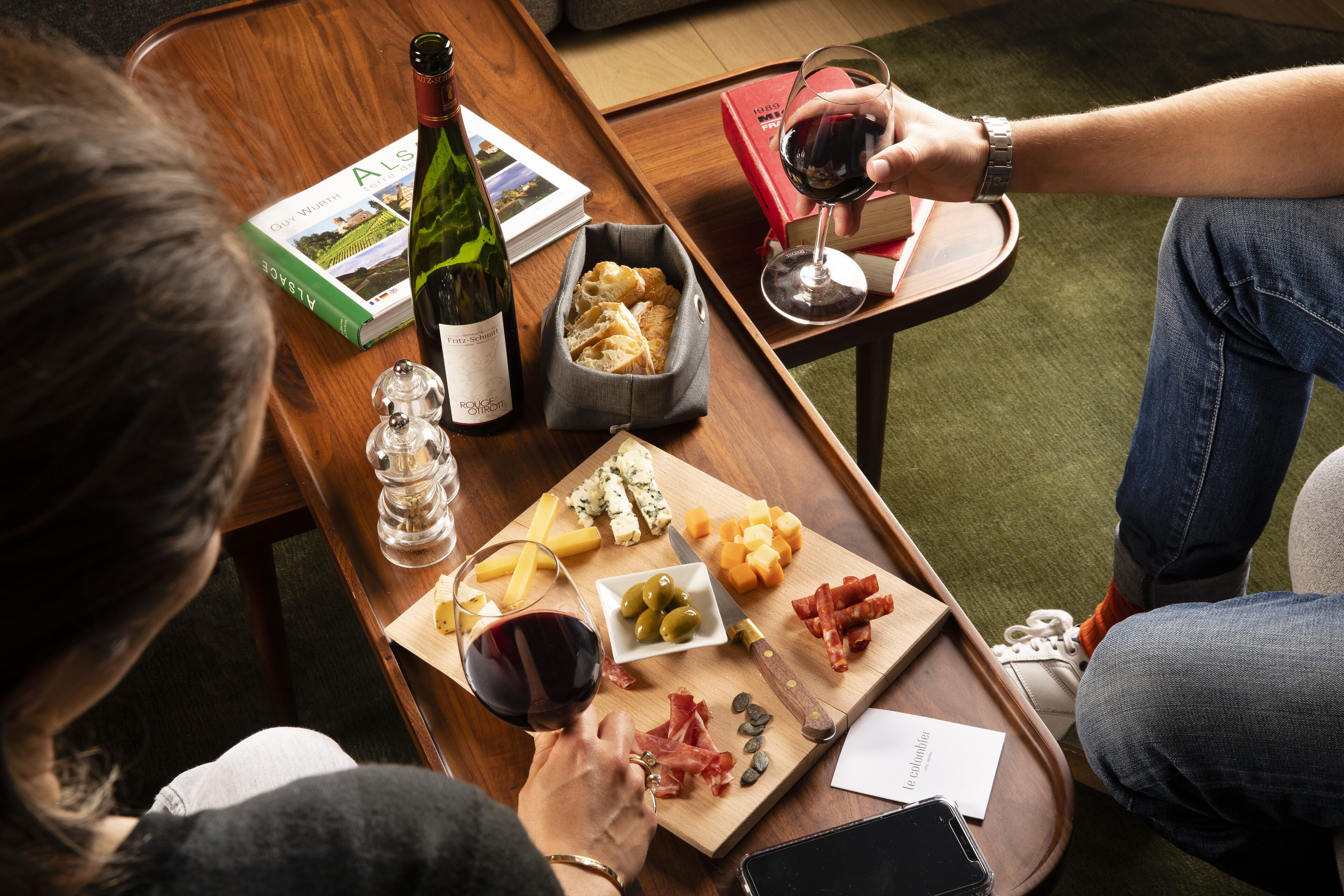 Een houten tafel met een wijnfles, een glas wijn, een boek, kaas, olijven en snacks.