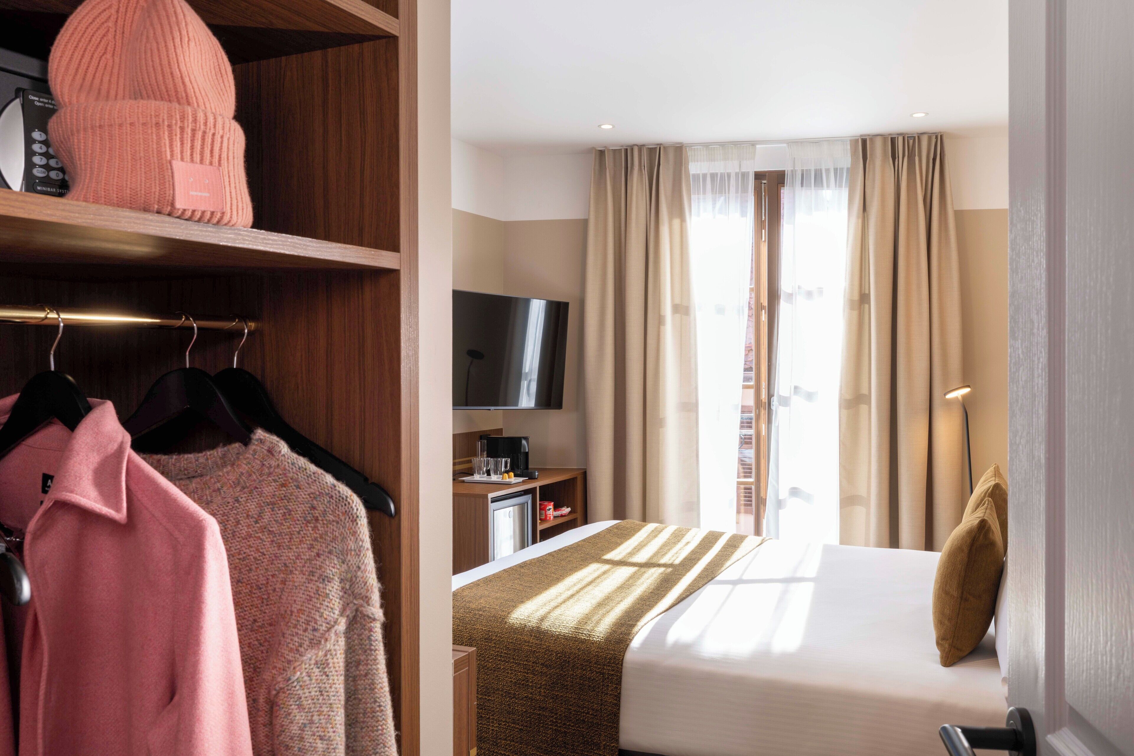 Une chambre d’hôtel avec un lit, une télévision, un bureau avec une machine à café et une armoire contenant des vêtements et un bonnet rose.