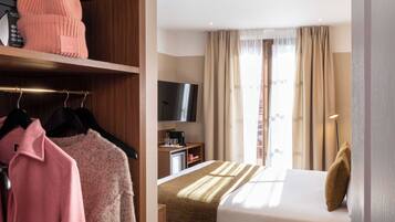 A hotel room with a bed, a TV, a desk with a coffee maker, and a wardrobe with clothes and a pink beanie.