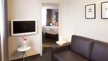 A hotel room with a large flat-screen TV, a sofa, a small round table with a vase of flowers, and a bed in the background.