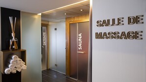 Un pasillo con una sala de masajes identificada como 'SALLE DE MASSAGE' y una sala de sauna señalizada como 'SAUNA'. Hay toallas apiladas en un estante y un difusor con palitos de caña.