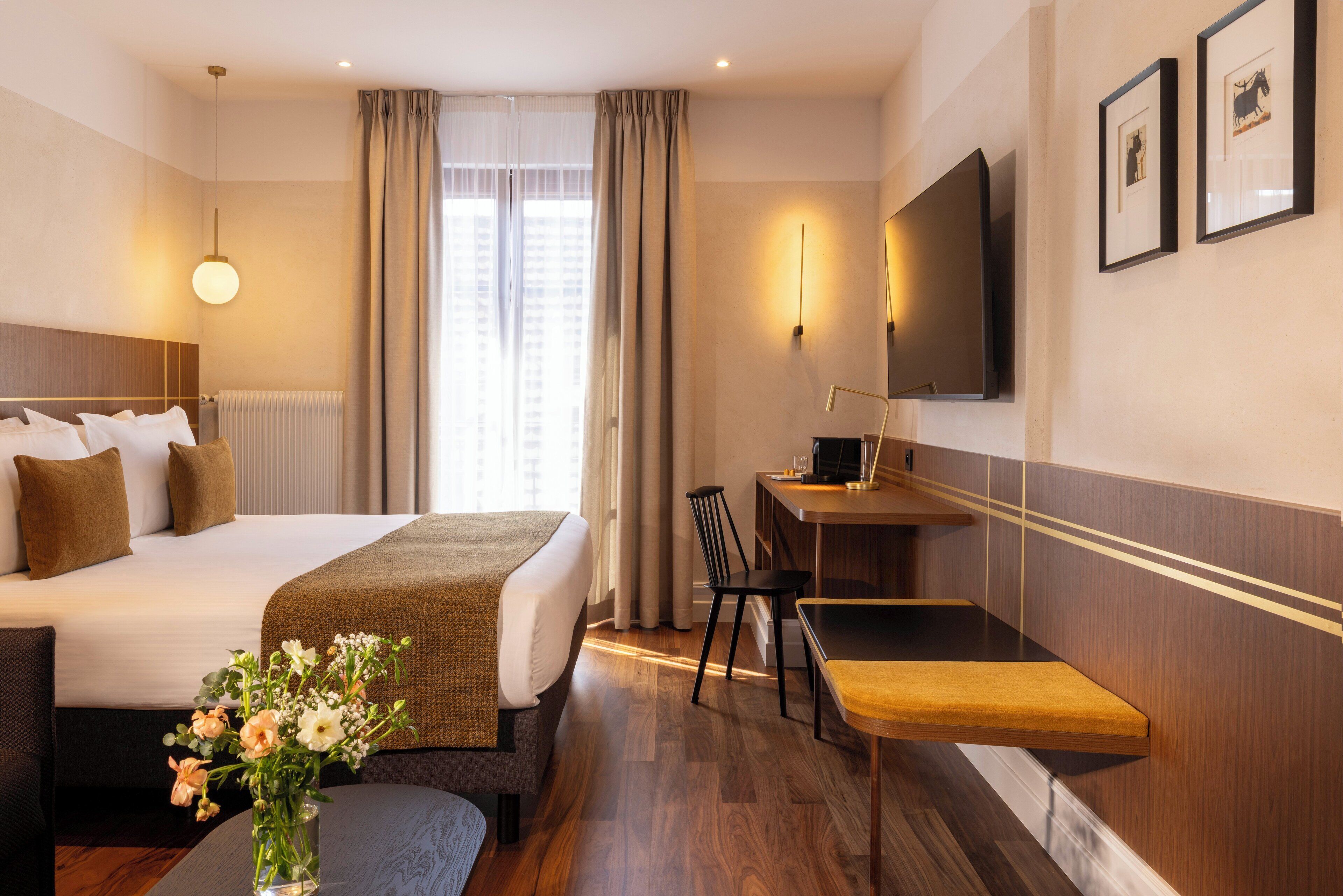 A hotel room with a bed, a desk, a chair, a small table, a vase with flowers, and a TV on the wall.