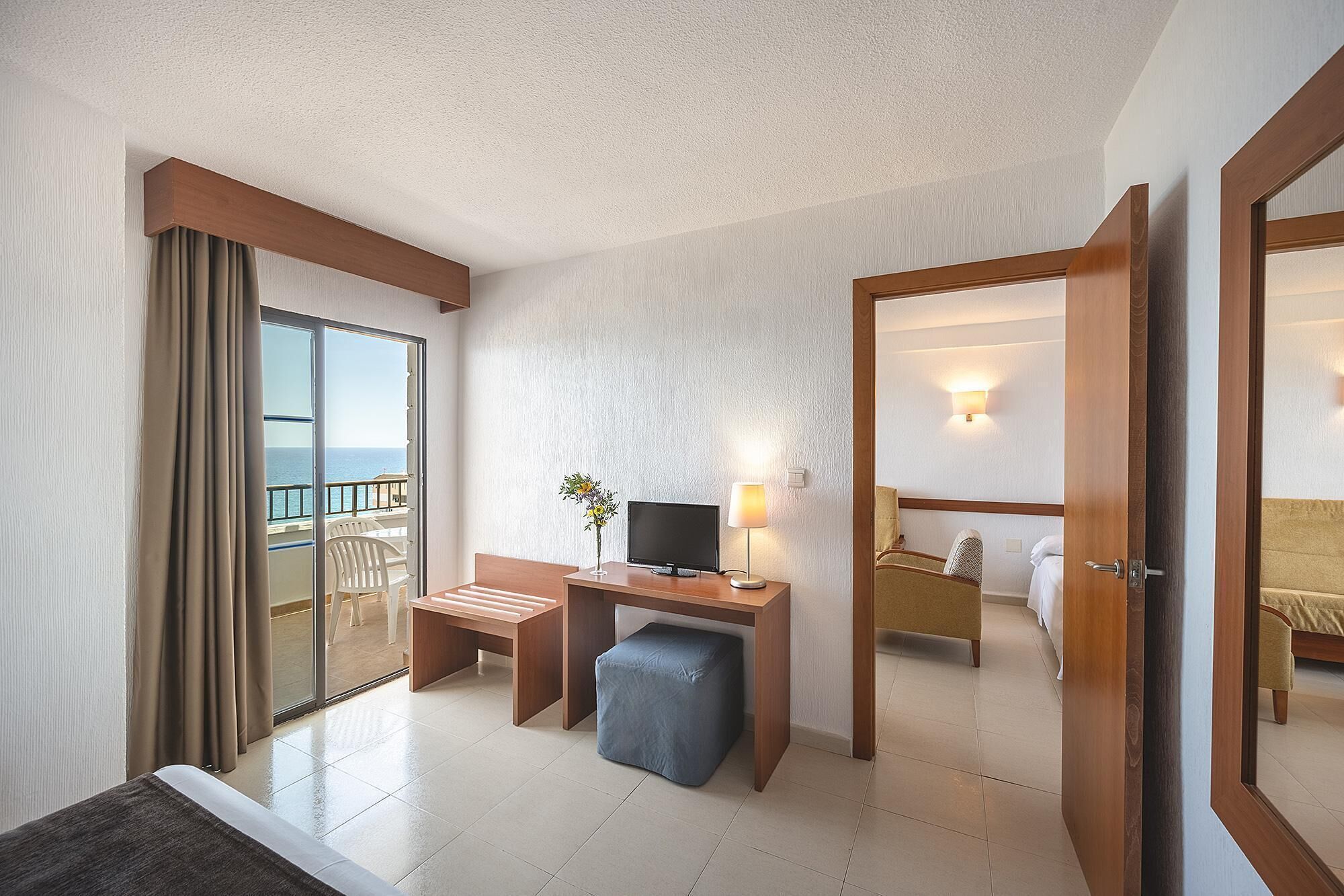 A hotel room with a balcony, a desk with a TV, a bedside table, a chair, and a bed.