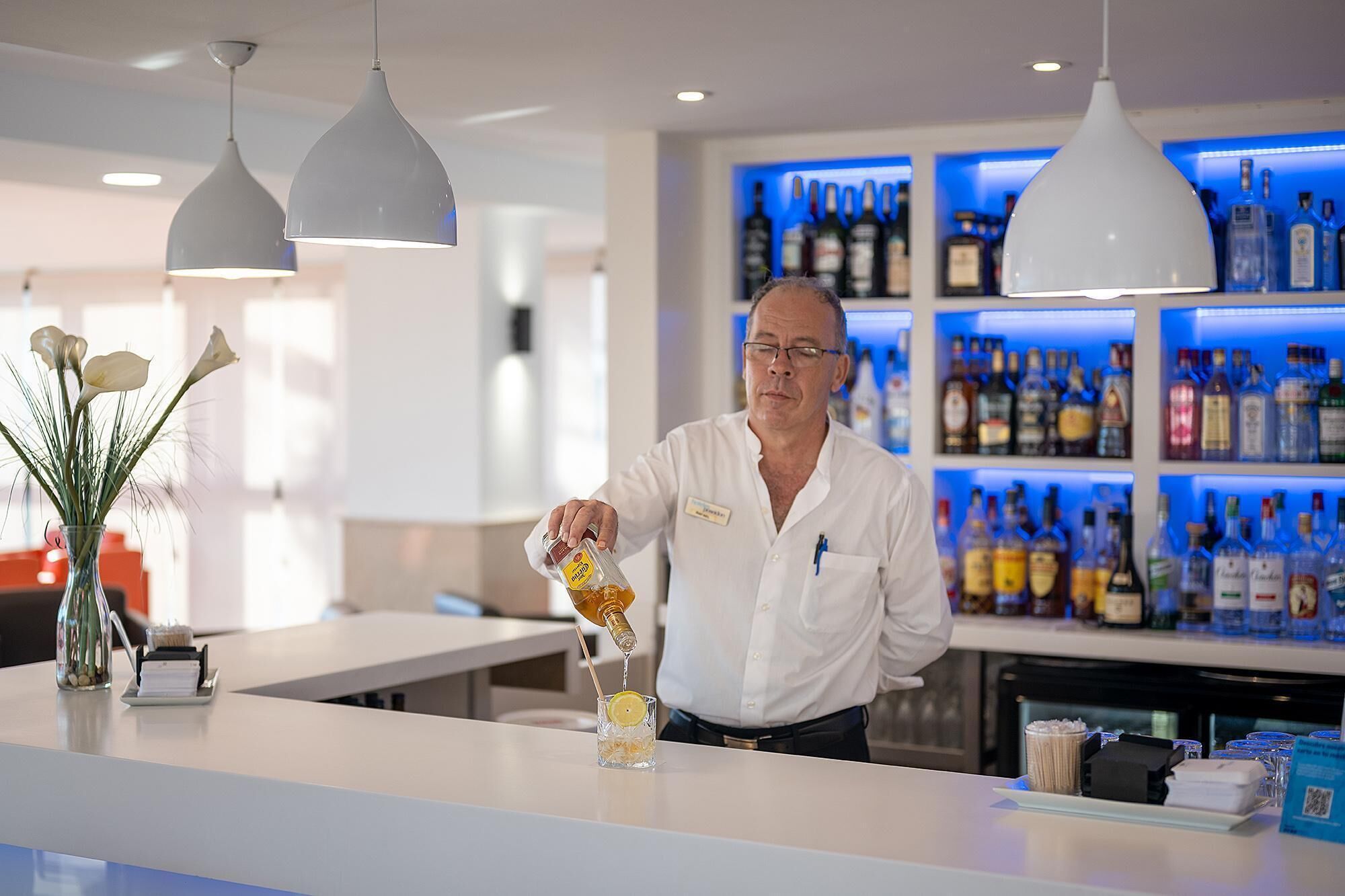 En bartender hælder en drik i et glas med et citronskive, i en moderne bar med velassorteret bar og hængende lamper.