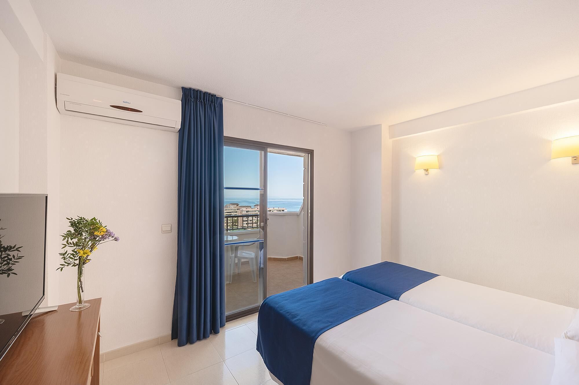 A hotel room with a bed, a TV, a desk, a vase with flowers, a balcony with a view, and a ceiling fan.