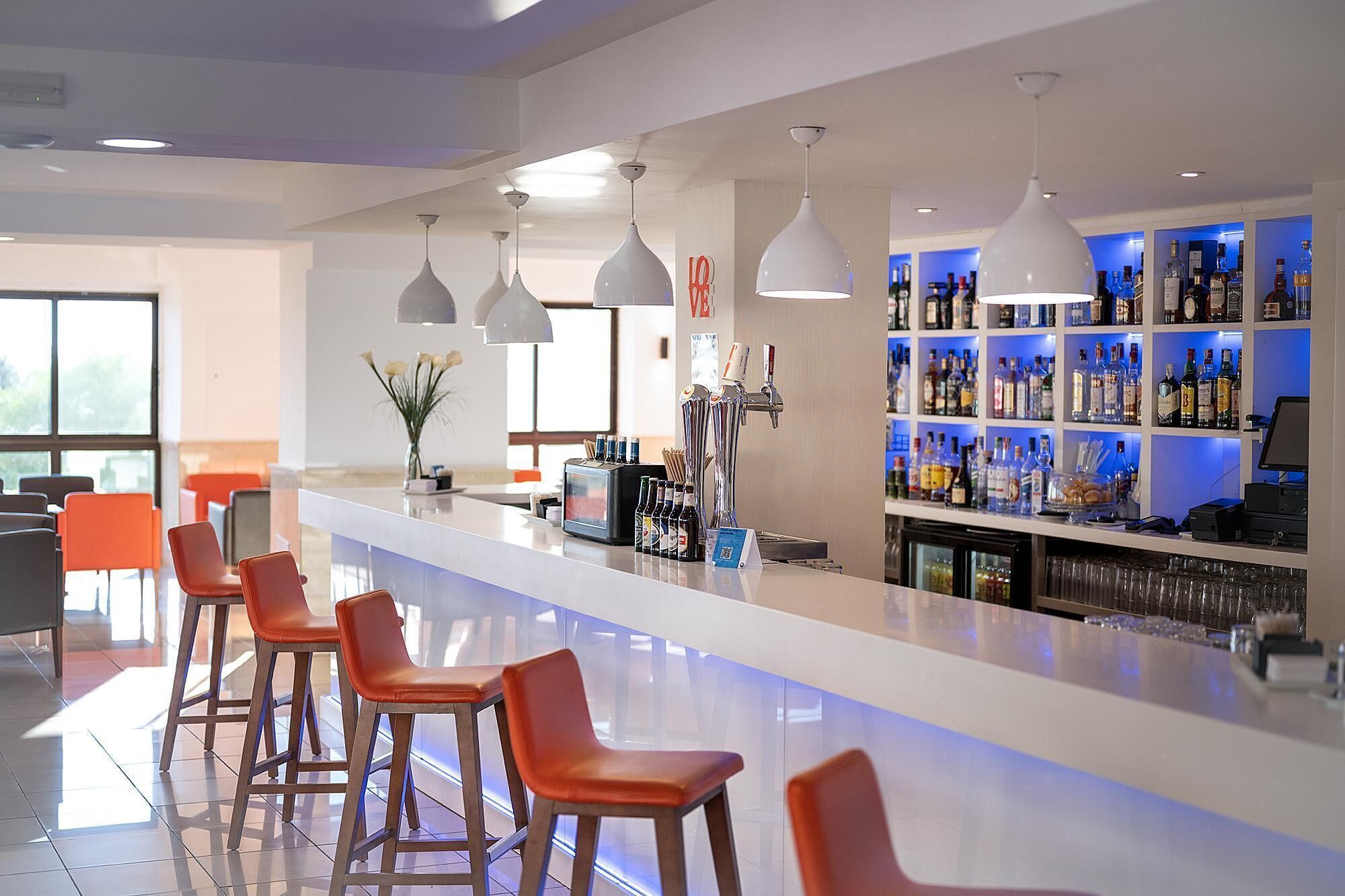 A modern bar area with a long counter, multiple bar stools, and a well-stocked liquor shelf.