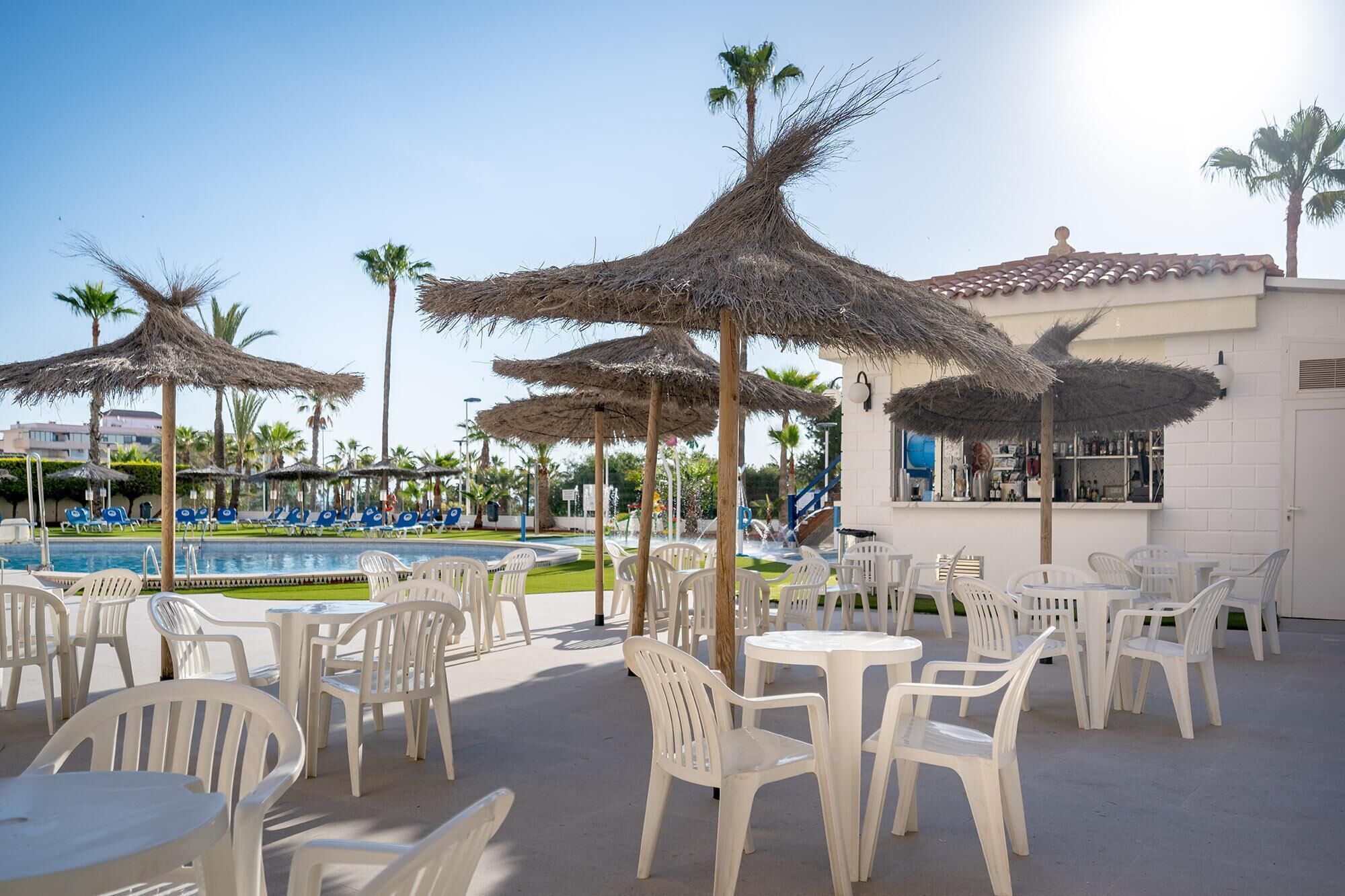 A poolside area with white plastic chairs and tables, palm trees, and a bar area with a white facade.