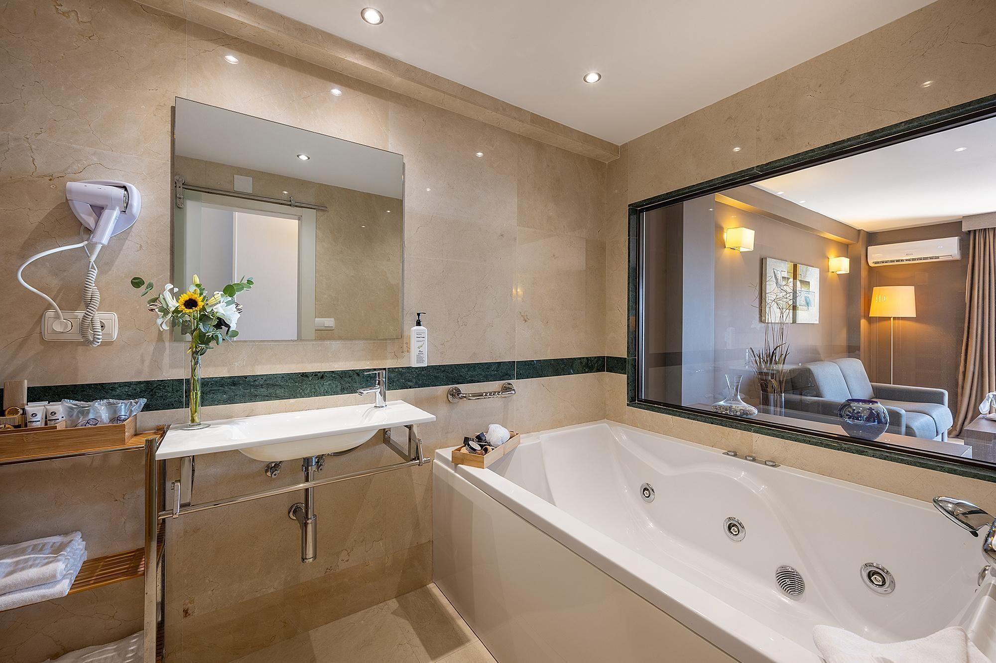 A modern bathroom with a large bathtub, a vanity with a mirror, a hairdryer, and a vase with flowers.
