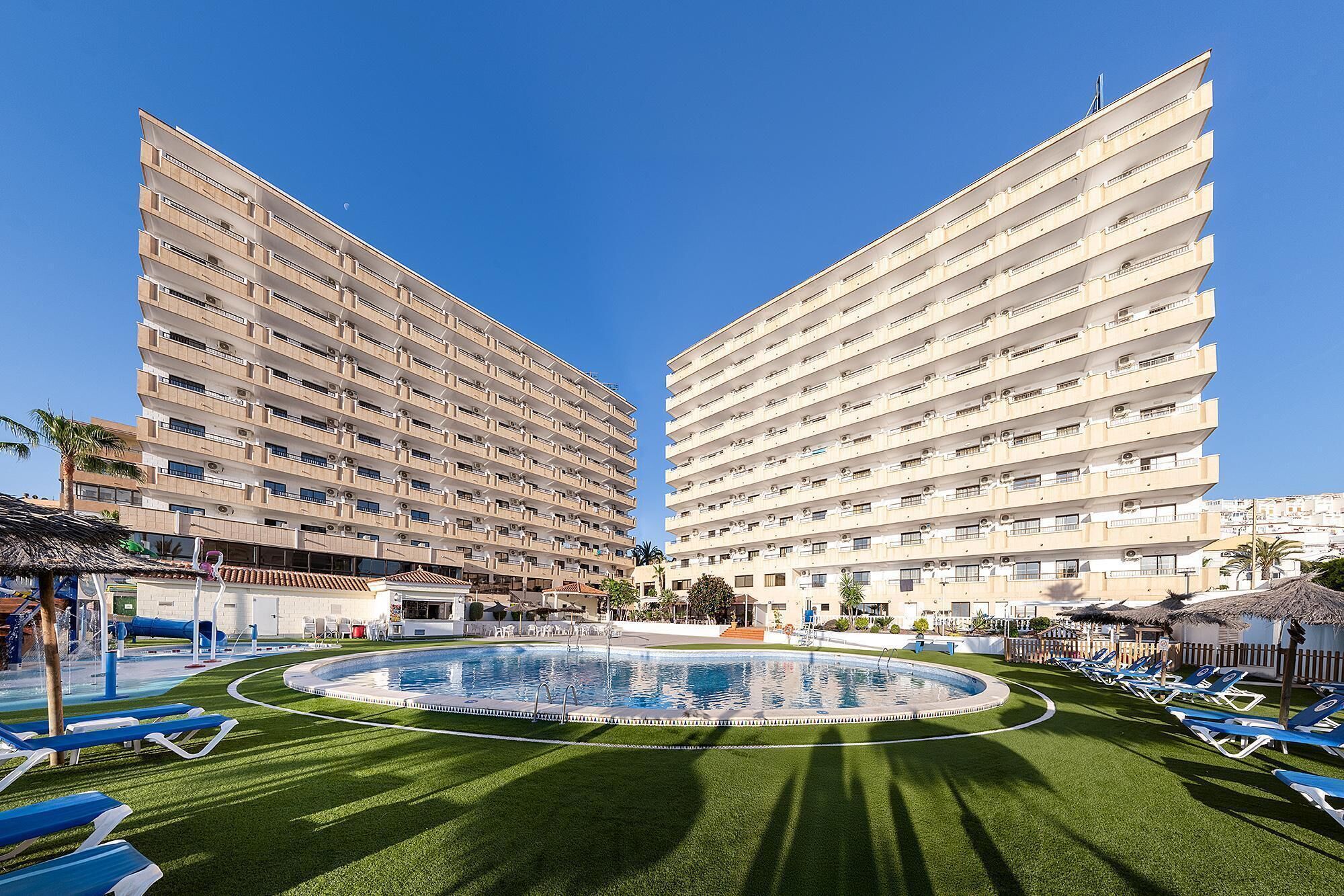 Ein Hotel mit einem zentral gelegenen Schwimmbecken, umgeben von Liegestühlen und einer gepflegten Rasenfläche.
