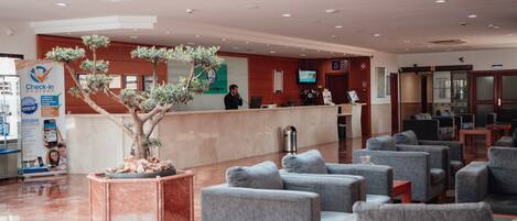 Un hall de hotel con un mostrador de recepción, un gran árbol en maceta y varias sillones grises.