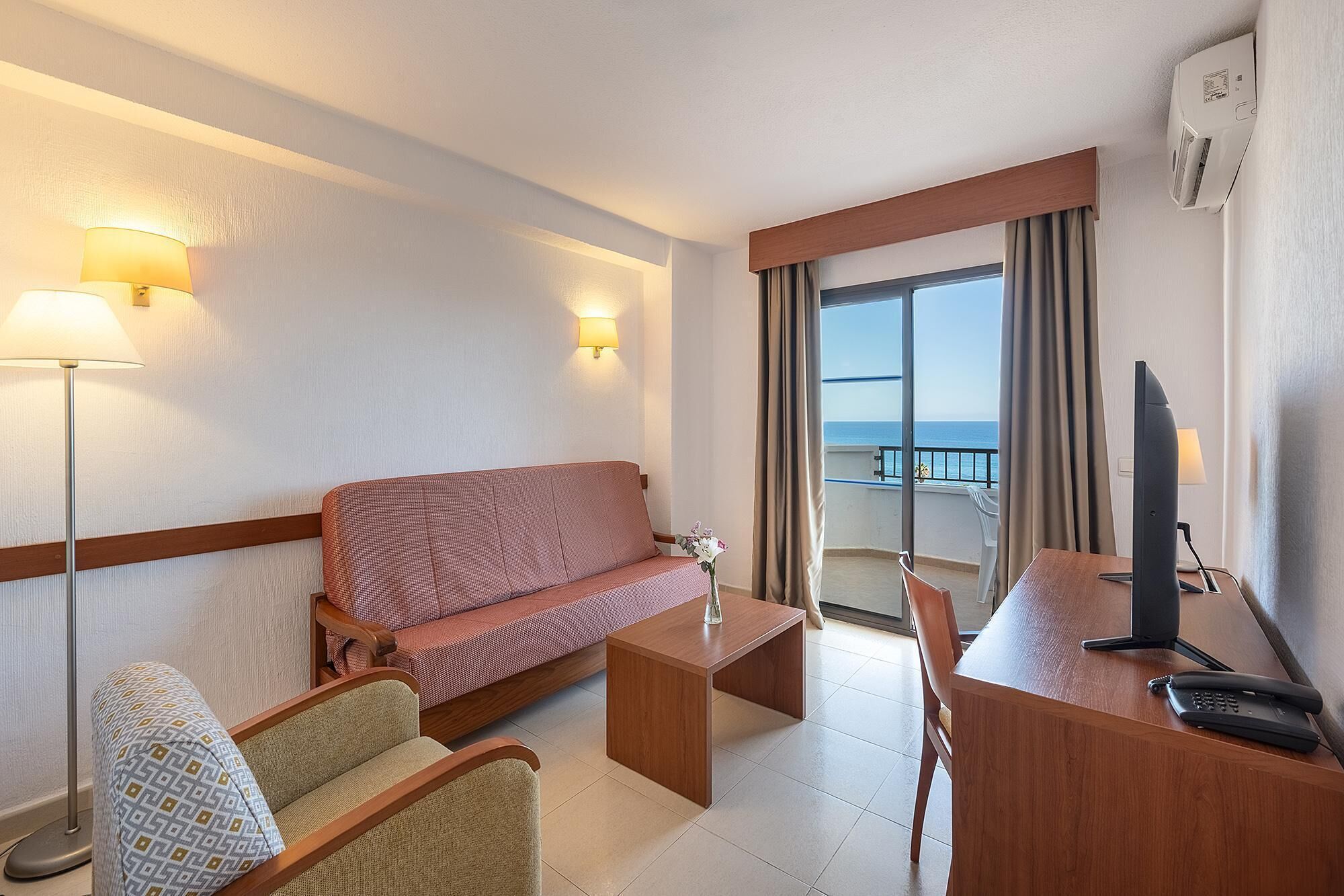A hotel room with a sofa, two armchairs, a desk with a computer, and a view of the sea.