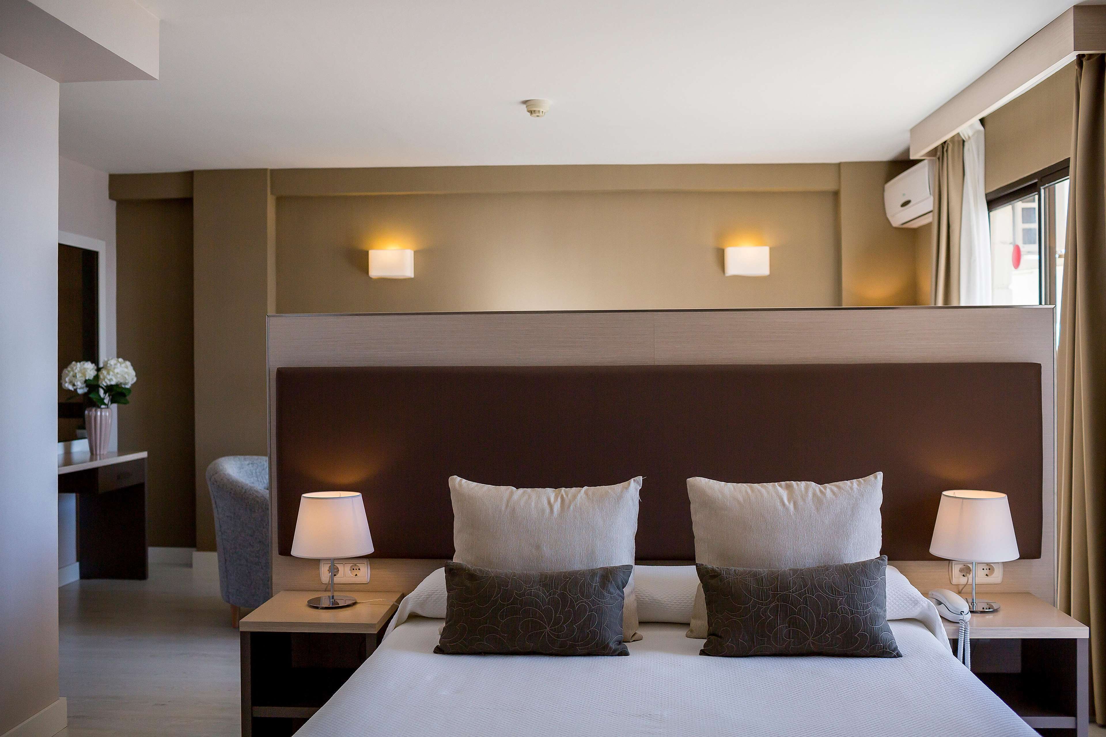 A hotel room with a large bed, two bedside lamps, a nightstand, a chair, and a wall-mounted air conditioner.