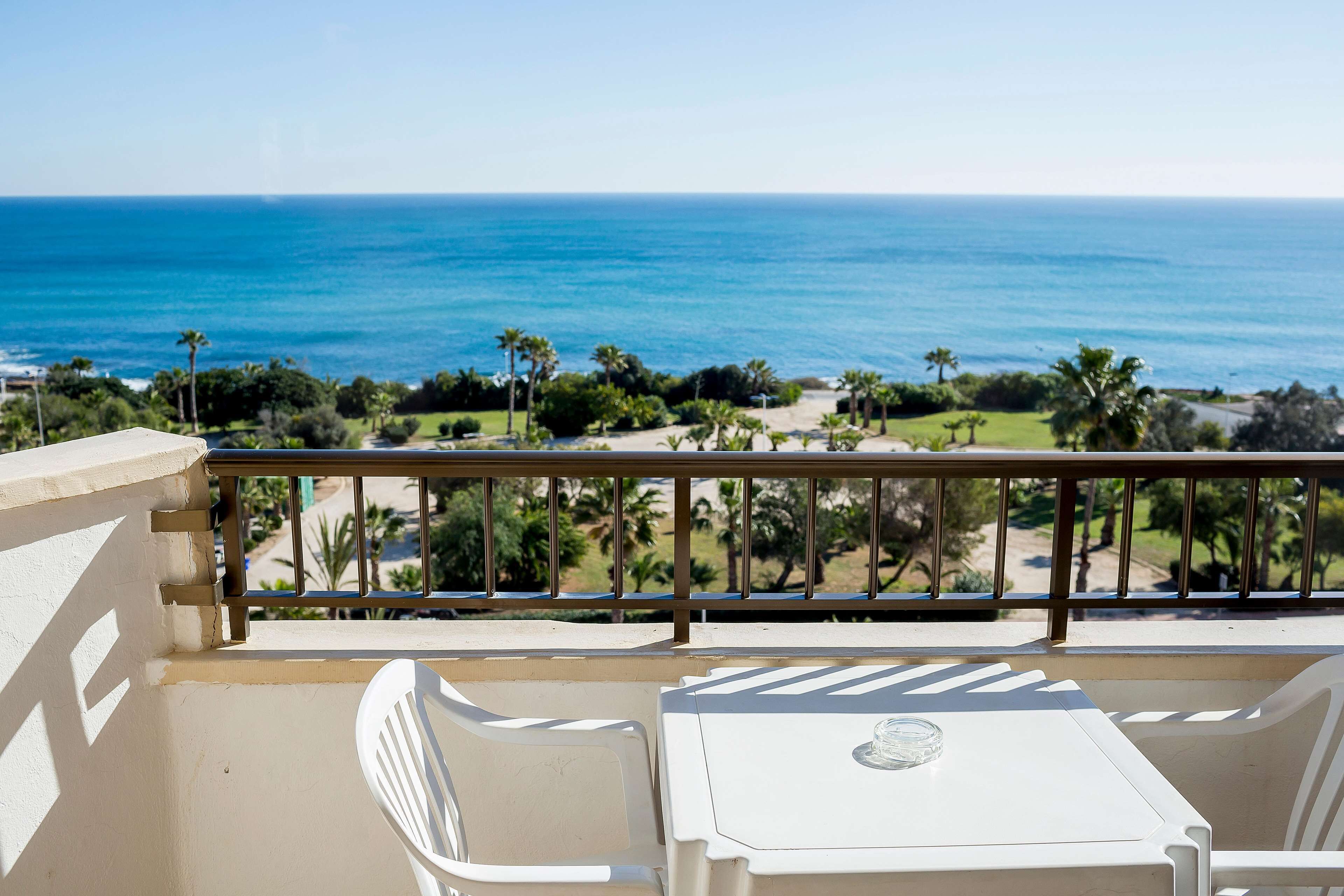 A balcony with a table and chairs, overlooking a beach with palm trees and the ocean.