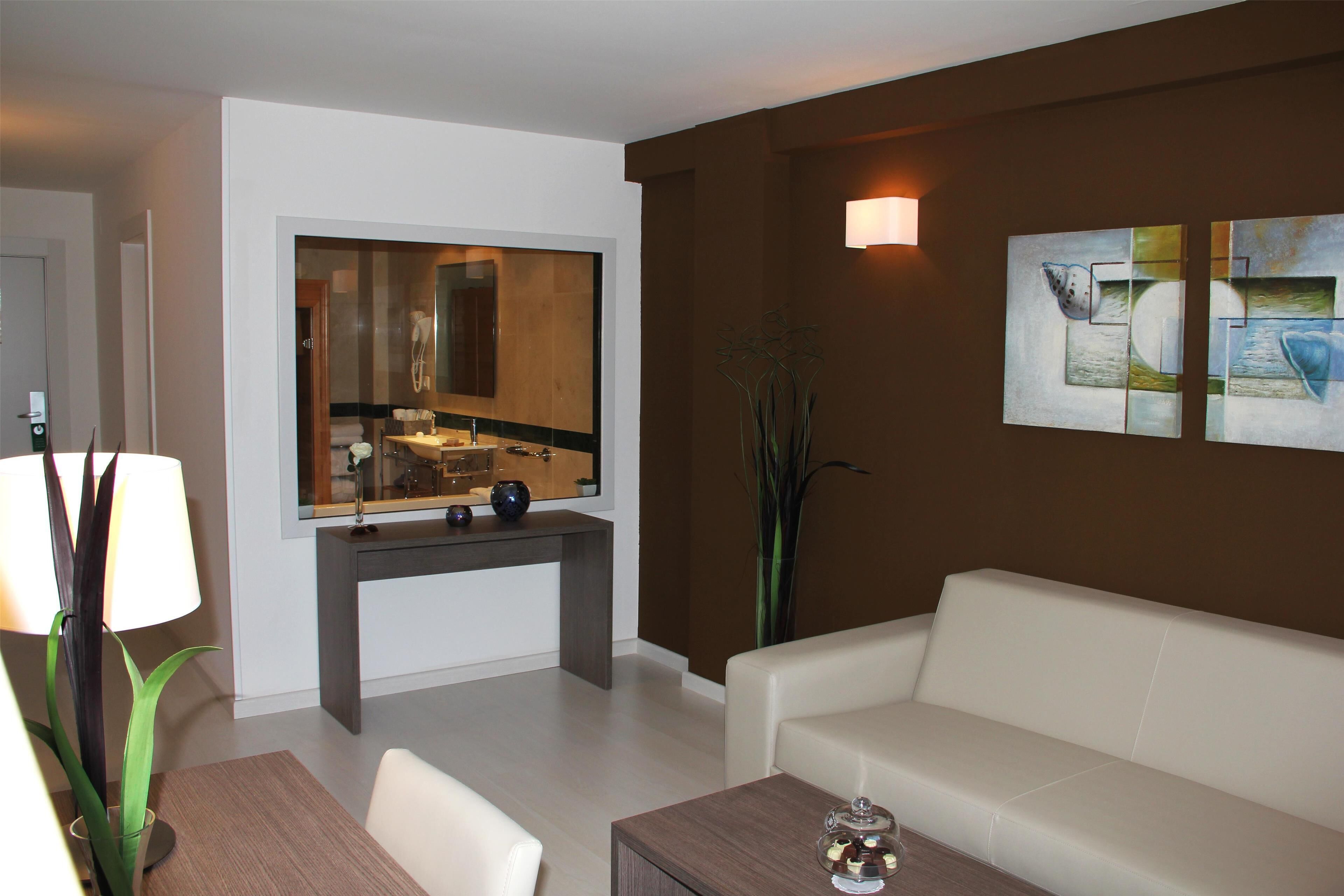 Ein modernes Wohnzimmer mit einem weißen Sofa, einem Holztisch, einem Esstisch mit Stühlen, einem großen Spiegel und abstrakter Wandkunst.