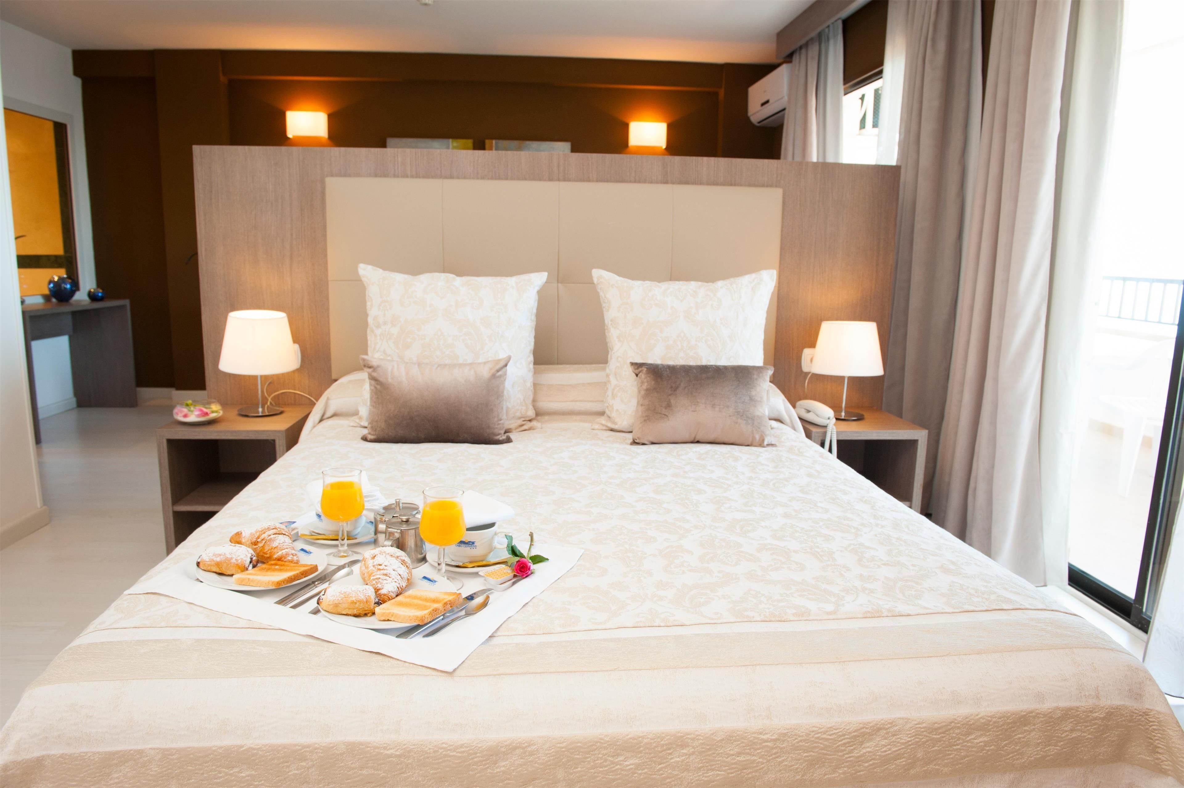 A hotel room with a large bed, two bedside lamps, a nightstand with a phone, and a breakfast tray with pastries and orange juice.