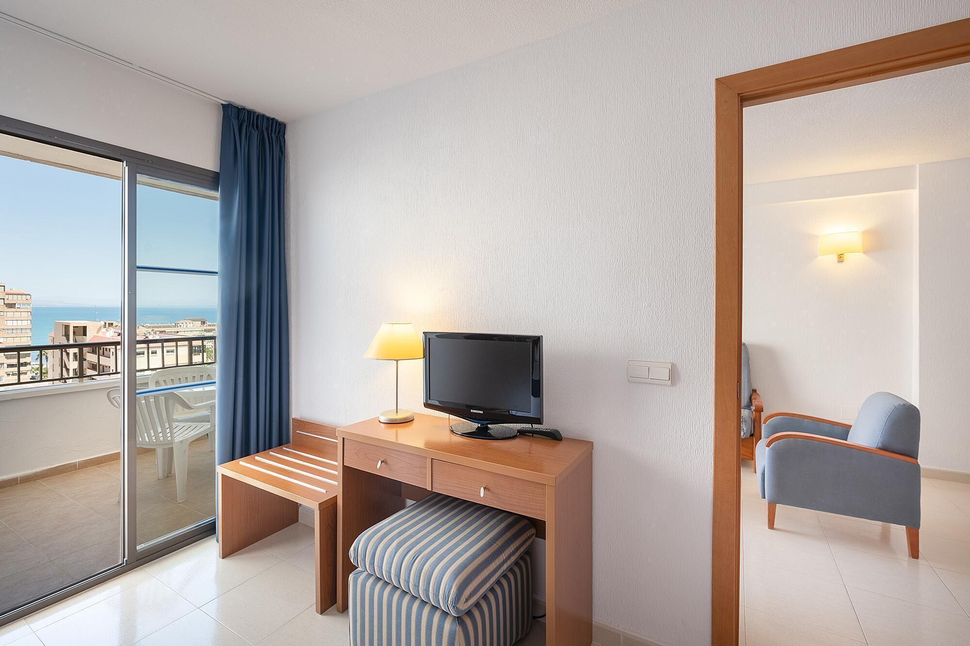 A hotel room with a balcony, a desk with a television, a chair, and a window with curtains.