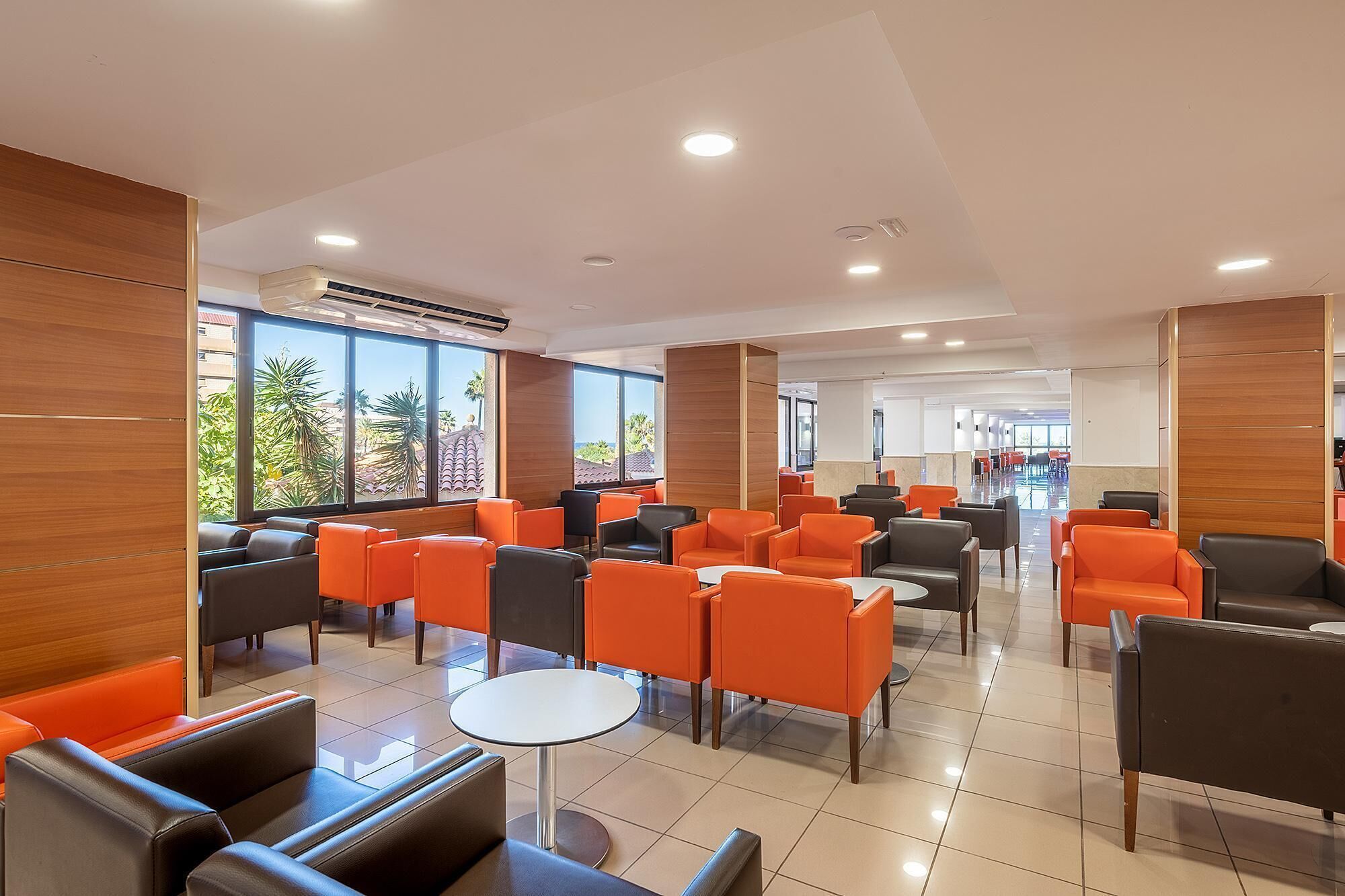 A modern indoor seating area with orange and black chairs, large windows, and a tiled floor.
